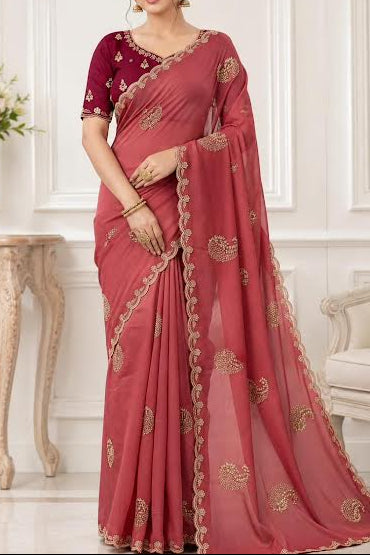 A woman models a dusty rose pink saree with gold embroidered motifs and a matching maroon blouse with gold embroidery, set against an elegant white paneled room background.