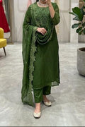 A woman takes a mirror selfie in a boutique wearing a green ethnic suit with matching dupatta and pants. The setting features a gray marble floor, a yellow accent chair, a potted plant, and a mannequin displaying a red dress in the background.