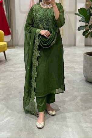 A woman takes a mirror selfie in a boutique wearing a green ethnic suit with matching dupatta and pants. The setting features a gray marble floor, a yellow accent chair, a potted plant, and a mannequin displaying a red dress in the background.