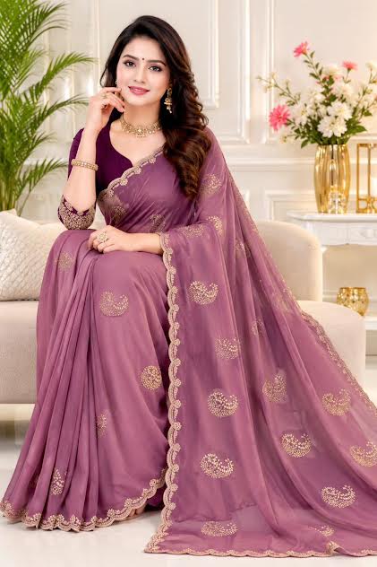 A woman seated on a sofa wearing a purple saree with gold floral embroidery and a matching blouse, in an indoor styled room with flowers and plants.