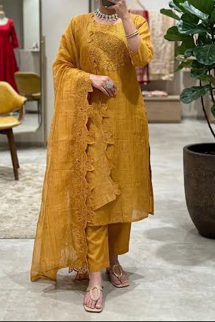 A woman takes a mirror selfie in a boutique wearing a mustard yellow traditional Indian outfit consisting of a kurta, pants, and a draped scarf. The boutique interior features a large mirror, a yellow chair, and a potted fiddle-leaf fig plant.