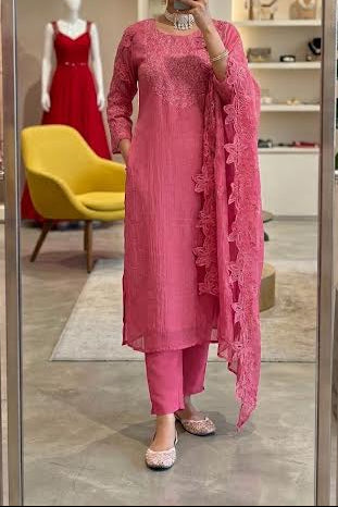 A woman takes a mirror selfie in a boutique wearing a pink traditional outfit with a matching embroidered dupatta. The setting features a yellow chair, a mannequin in a red dress, and a large potted plant.
