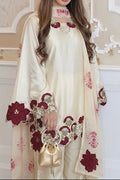 A woman models a cream-colored traditional outfit with deep red and pink floral embroidery, holding a matching cream handbag with a gold handle, set against an ornate beige and white wall.