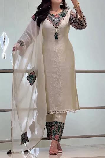 A woman takes a mirror selfie wearing a traditional Indian salwar kameez outfit. The set features a cream-colored embroidered kameez tunic with a green and gold embroidered neckline, patterned sleeves, matching pants, and a sheer white dupatta with green and gold embroidered corner accents.