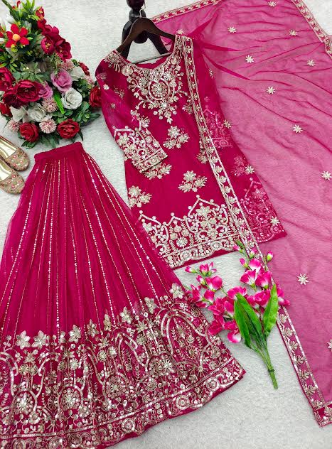 A bright pink traditional outfit set with heavy gold embroidery, including a kurta, matching skirt, and dupatta, arranged on a light gray surface with decorative flowers.