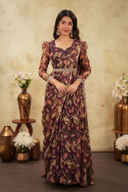 A woman stands indoors wearing a long-sleeved, floor-length floral dress in deep purple and beige tones, with a patterned belt at the waist. The background features a textured beige wall with decorative brass vases and flowers.