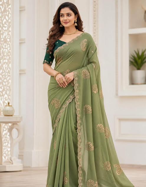 A woman modeling a sage green georgette saree with gold floral embroidery and a matching green embroidered blouse, set against a bright, white-toned indoor background.