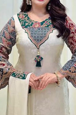 A woman wearing a cream-colored traditional dress with ornate multicolored embroidered sleeves and a detailed neckline, standing against a light-colored paneled wall.
